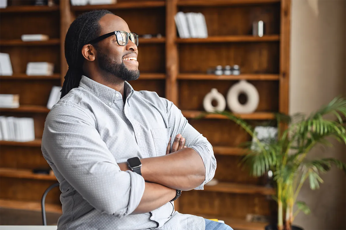 Black man standing with folded arms beside bookshelves